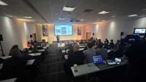 A wide elevated view of a filled corporate auditorium during a presentation, showing attendees seated at long desks facing a stage projector screen and a rear video production station.