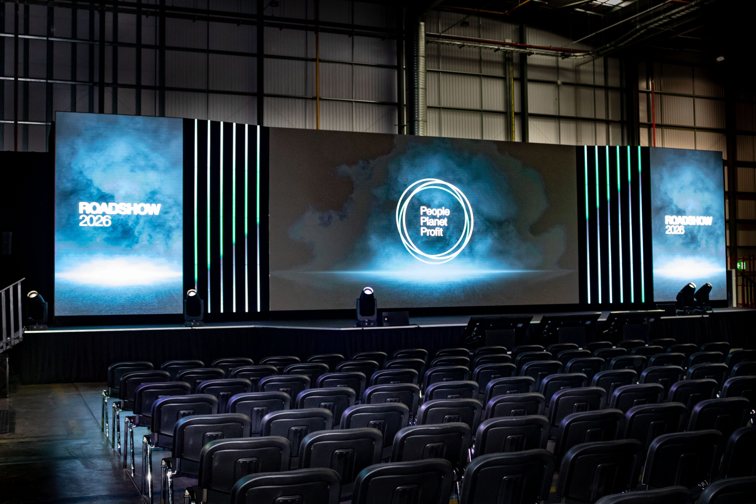 Wide-format modular LED stage wall displaying 'Roadshow 2026' and 'People Planet Profit' graphics with vertical light accents and empty theatre seating in front.