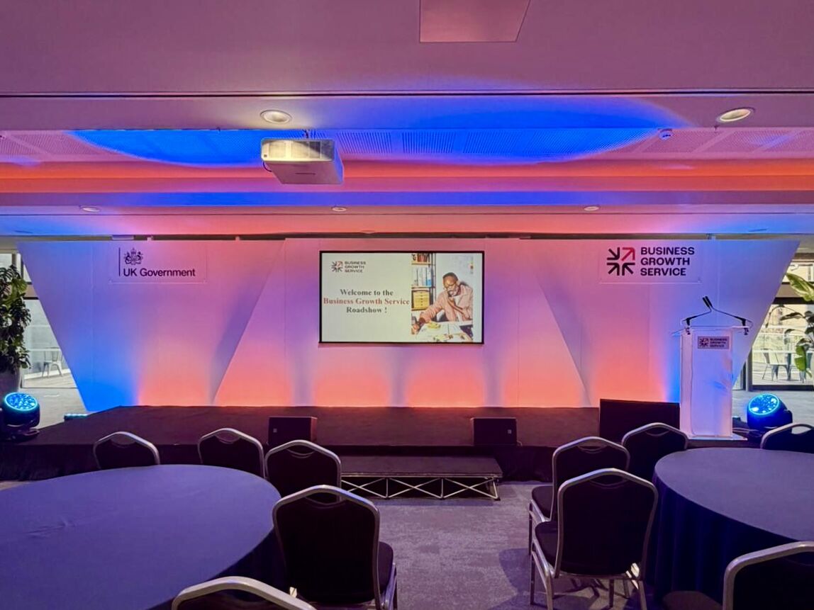 Branded corporate event stage for the UK Government Business Growth Service roadshow, with illuminated backdrop, presentation screen, lectern and seated audience tables.